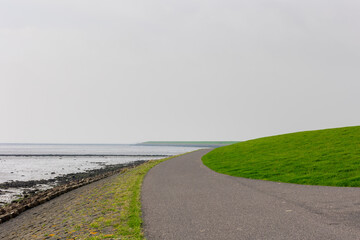 Bicycle lane and walkway, Green grass meadow on the dyke under cloudy sky, Dike between polder land and north sea with fog or mist in the morning, Dutch Wadden Sea island, Terschelling, Netherlands.