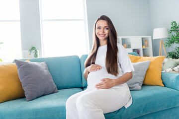 Photo of tender adorable pregnant girl sitting on sofa touching belly feeling baby waiting for childbirth