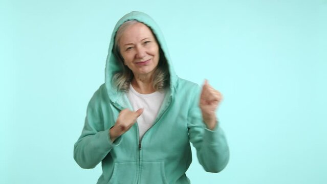 A happy senior lady dances in a casual hoodie, her face beaming with joy and energy, bringing a sense of fun and vitality on a light teal background. Camera 8K RAW. 