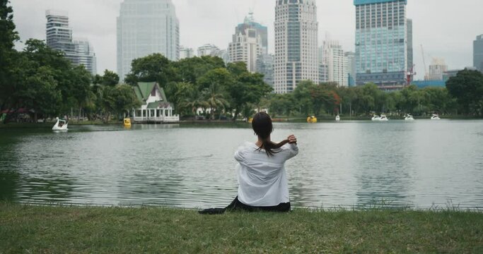 Woman sits on grass in city park Bangkok skyscrapers background lakeside back view Tranquility in urban nature. Tranquility fusion of city and greenery Tranquility amidst Thailand's flora.