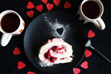 A piece of meringue roll on a black plate next to two cups of tea on a dark background