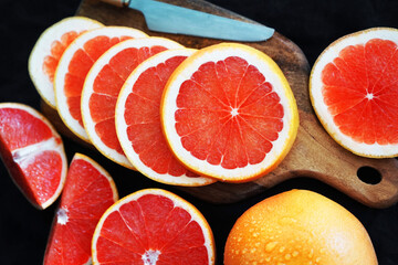 Sliced ​​and whole grapefruit on a wooden board next to a knife on a dark background