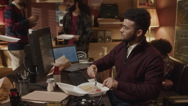 Young Biracial male programmer having lunch together with mates and colleagues in handmade apartment office