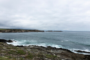 Fototapeta na wymiar A rugged coastline with turbulent waves under a cloudy sky, surrounded by distant cliffs and greenery