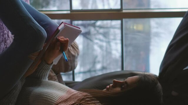 Vertical video. Diary leisure. Writing inspiration. Young pensive creative woman with stories notebook at cozy home with winter window view.