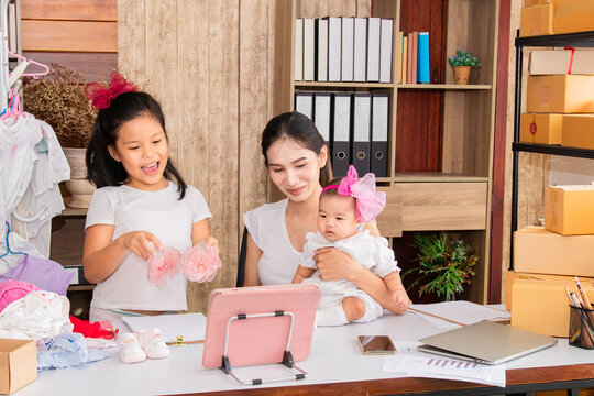Asian parent working while looking after a young baby or toddler, single mother businesswoman working from home multitask nursing infant using technology modern laptop online selling children clothes