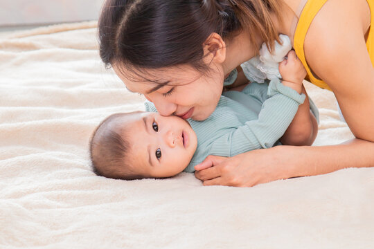 Selective focus of Asian young mother kissing baby on bed, Beautiful woman hugging toddler infant lying down together on soft fur. Cute newborn child playing with mom. family motherhood health care