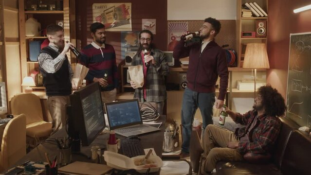 Team of multiethnic mates celebrating software startup launch together in old-fashioned vintage-style apartment, cheering and toasting with glass beer bottles