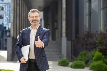 Smiling senior businessman in smart suit standing outdoors with a laptop, giving a thumbs up in...