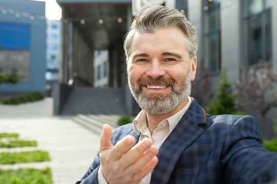 Portrait of a professional senior businessman with a welcoming gesture, standing confidently outside a modern office building.