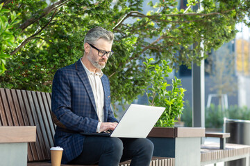 Mature professional financier in suit focused on work with laptop on bench outside. Urban business lifestyle captured in natural daylight.