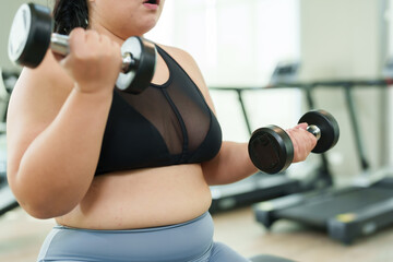 Fototapeta na wymiar Overweight Asian woman exercise in gym, Focused in activewear, exuding confidence and dedication in workout. Concentrated female doing arm exercises with weights, showing commitment to fitness against