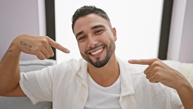 Cheerful young arab man, sitting comfortably on his sofa at home, proudly showing off his pristine teeth with a beaming smile. pointing to his dental health, he's a perfect case of dazzling oral care.