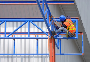 Welder with safety equipment is welding metal roof beam on top of factory building outline structure in construction site, low angle view
