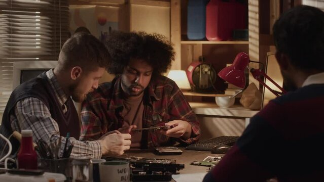 Waist up of diverse male team of soldering technician having discussion while repairing motherboard on desk in dark workshop with yellow dimmed light