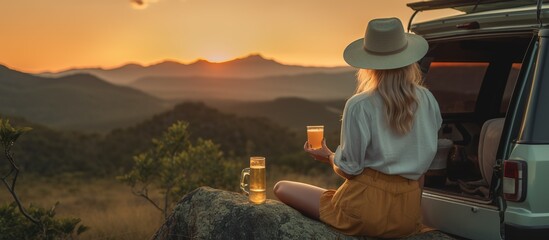 seen from behind Beautiful young female traveler wearing a hat enjoying the sunset in the mountains while next to the car, holding a cup