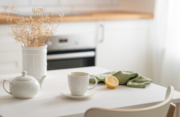 A cup of tea with lemon and a teapot on a white table with dry bouquet.