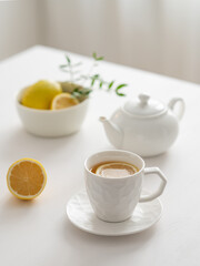 A cup of tea with lemon and a teapot on a white table against the background of a kitchen window.