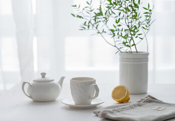 A cup of tea with lemon, a teapot and green branch on a white table against the background of a...