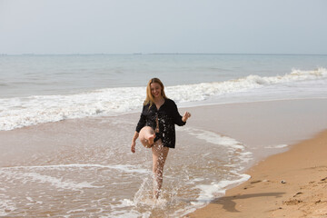 Young, pretty blonde woman walks along the shore of the beach wearing a black shirt and black...