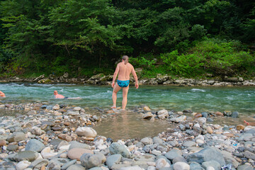 Man with Long Hair Swimming in Scenic Mountain River with Trees and Stones in Background