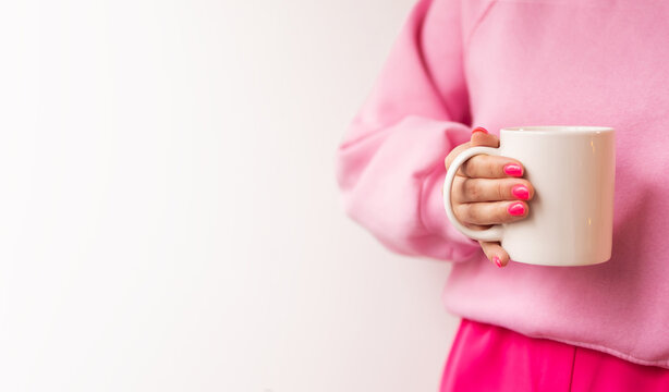Female hands pink manicure holding white mug mockup blank space to copy your advertising text message, mockup. Girl in pink sweatshirt, fuccia pants, gray wall, mockup, close-up, advertising content