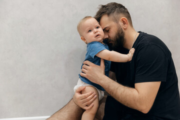 Father hugs baby boy and calms and wipes away tears, young man kisses his son on head in living...
