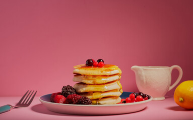 Stack of fresh pancakes with mixed berries and honey running down the sides on a pink background.