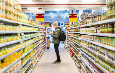 Male customer perusing food items on supermarket shelves in grocery store. Man looks at price and choose organic food.