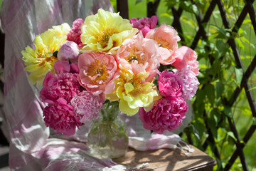 Bouquet of yellow, pink and coral peonies in a vase on a chair in the garden by the pergola against the backdrop of green grass. Colorful postcard