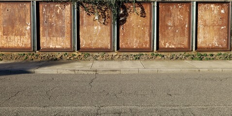 Rusty metal boards with scraped street billboards and ivy on top, at the roadside . Concrete...