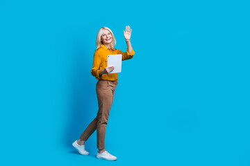 Full length photo of blonde lovely senior lady hold tablet wave hand dressed stylish yellow satin garment isolated on blue color background