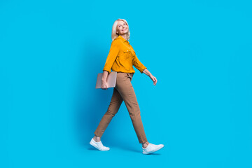 Fototapeta na wymiar Full body photo of blonde pensioner woman hold device confident walk dressed stylish yellow satin clothes isolated on blue color background