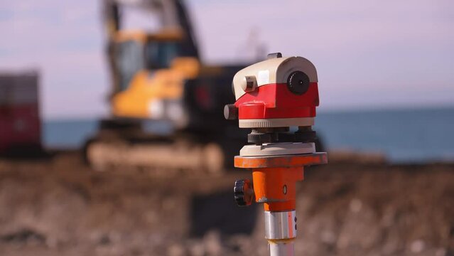 Close-up image of optical level or surveying instrument used to measure angles and distances on construction site. In background, construction worker using crane to lift materials and bulldozer . sea