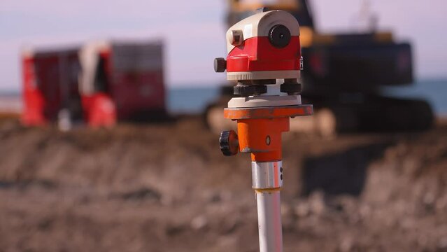 Close-up image of optical level or surveying instrument used to measure angles and distances on construction site. In background, construction worker using crane to lift materials and bulldozer . sea