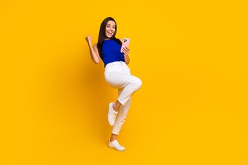 Fototapeta na wymiar Full length photo of positive lucky woman wear blue top rising fist winning game modern gadget empty space isolated yellow color background