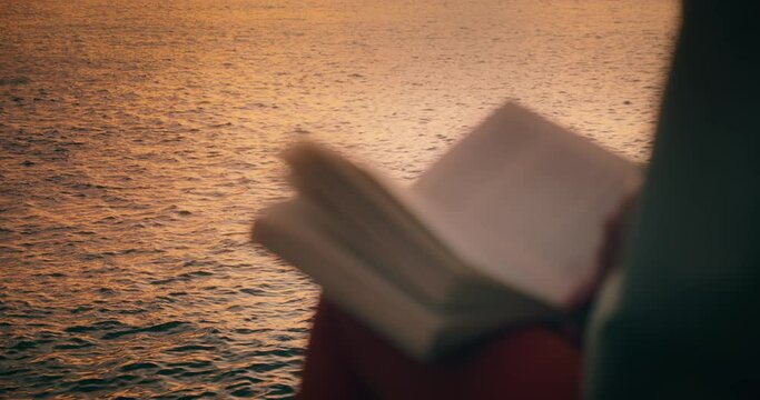 Girl leafing through the pages of a book on rocky beach with ocean view at sunset.