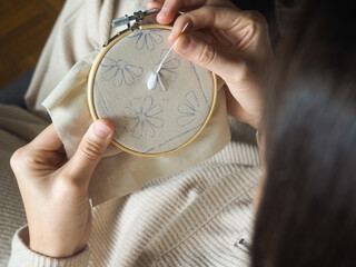 Detail view of two hands embroidering in round bamboo frame. An unrecognizable person is embroidering some white flowers on a beige fabric. The woman is in lounge clothes, comfortable, on her sofa.