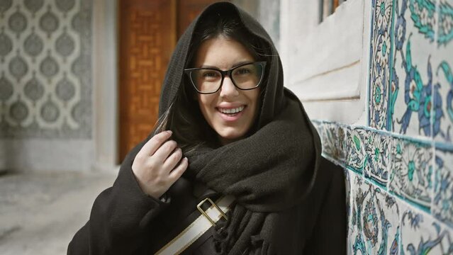 A beautiful young woman in glasses, smiles warmly leaning on an ottoman-era tiled wall in istanbul, showcasing culture and tourism.