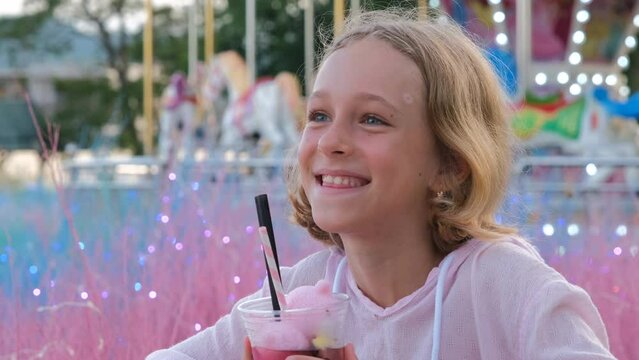 Happy little girl drinking lemonade over vintage horse carousel, slow motion. Enjoying evening in a funfair or an amusement park 