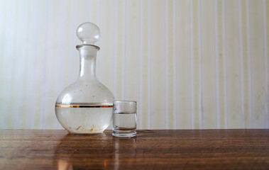 Glass dirty retro decanter with vodka and one shot located on wooden table in apartment with old...