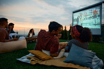 Happy couple eating popcorn while watching movie with friends on patio in evening.