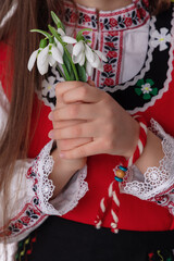Bulgarian woman in ethnic folklore embroidery dress nosia, yarn bracelet martenitsa, and spring bouquet snowdrops symbol of Bulgaria culture and spirit  - 725810949