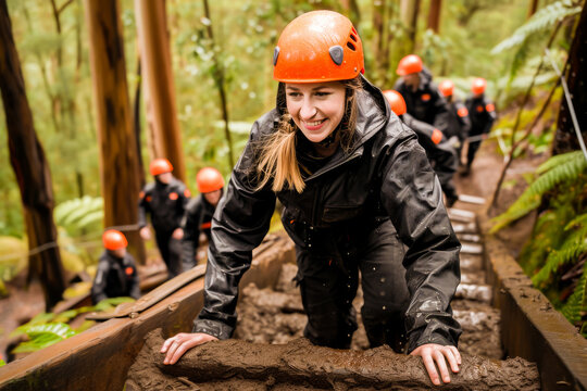 Smiling woman in a helmet climbing through a muddy obstacle in a forest adventure with a group of people in the background.