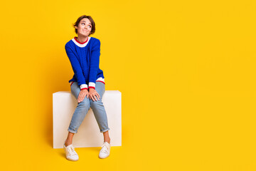 Full length photo of doubtful unsure lady dressed knitted sweater sitting cube looking empty space isolated yellow color background - obrazy, fototapety, plakaty