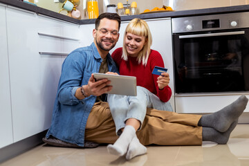 Happy couple sitting on the kitchen floor and ordering something online with their credit card