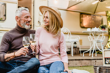 Senior couple on a date during picnic looking at each other with love drinking wine in the porch of their camper van. Spouses celebrating anniversary on a trip holiday by trailer motor home - obrazy, fototapety, plakaty