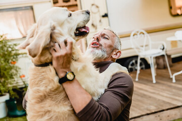 Portrait of happy mature caucasian man traveler pet owner with tattoo holding his labrador dog golden retriever in his hands in motorhome yard trailer camper van