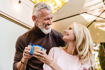 Handsome grey-haired mature man husband giving his beautiful blonde wife a present in their garden,...