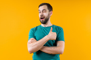 Studio portrait of confident young man posing over bright colored yellow background winking and holding thumb up - 725860944